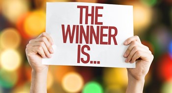 Who’s a Good Board? Two hands holding a sign reading THE WINNER IS...in front of a blurred multicolored background