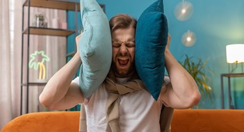 Respecting Rules & Rights Repair work at neighbours. Irritated young man relaxing on couch cover ears with pillows annoyed by noisy neighbors suffer from headache wish silence. Thin walls at home flat without sound insulation