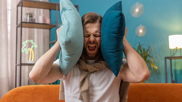 Repair work at neighbours. Irritated young man relaxing on couch cover ears with pillows annoyed by noisy neighbors suffer from headache wish silence. Thin walls at home flat without sound insulation
