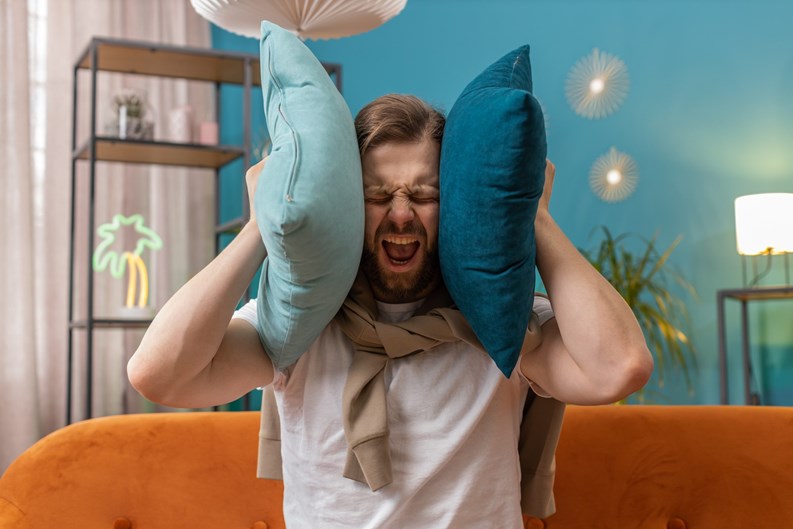 Repair work at neighbours. Irritated young man relaxing on couch cover ears with pillows annoyed by noisy neighbors suffer from headache wish silence. Thin walls at home flat without sound insulation