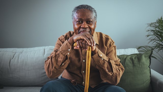 Portrait of happy senior man smiling at home while holding walking cane. Old man relaxing on sofa and looking at camera. Portrait of elderly man enjoying retirement.