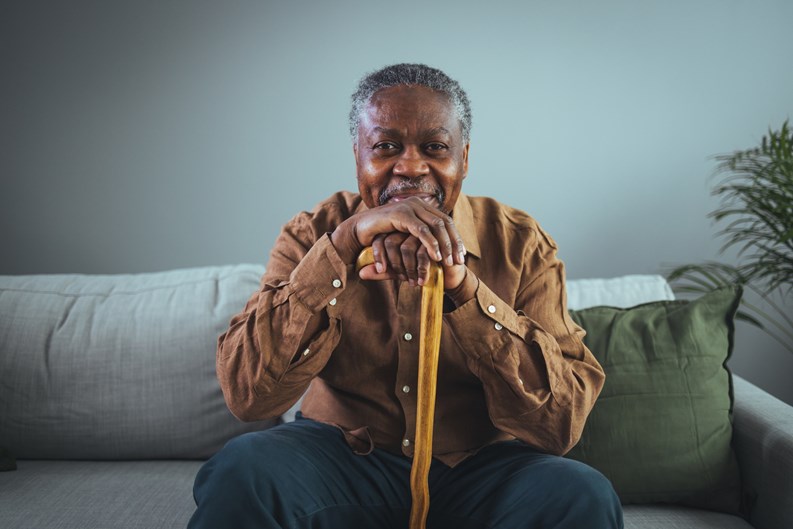 Portrait of happy senior man smiling at home while holding walking cane. Old man relaxing on sofa and looking at camera. Portrait of elderly man enjoying retirement.