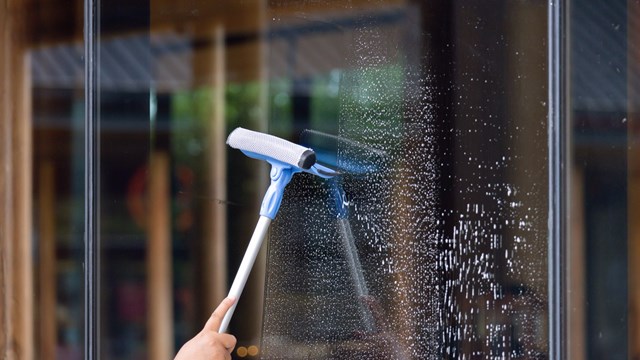 A person is cleaning a window with a squeegee. The window is covered in streaks and the person is using a blue and white squeegee to clean it