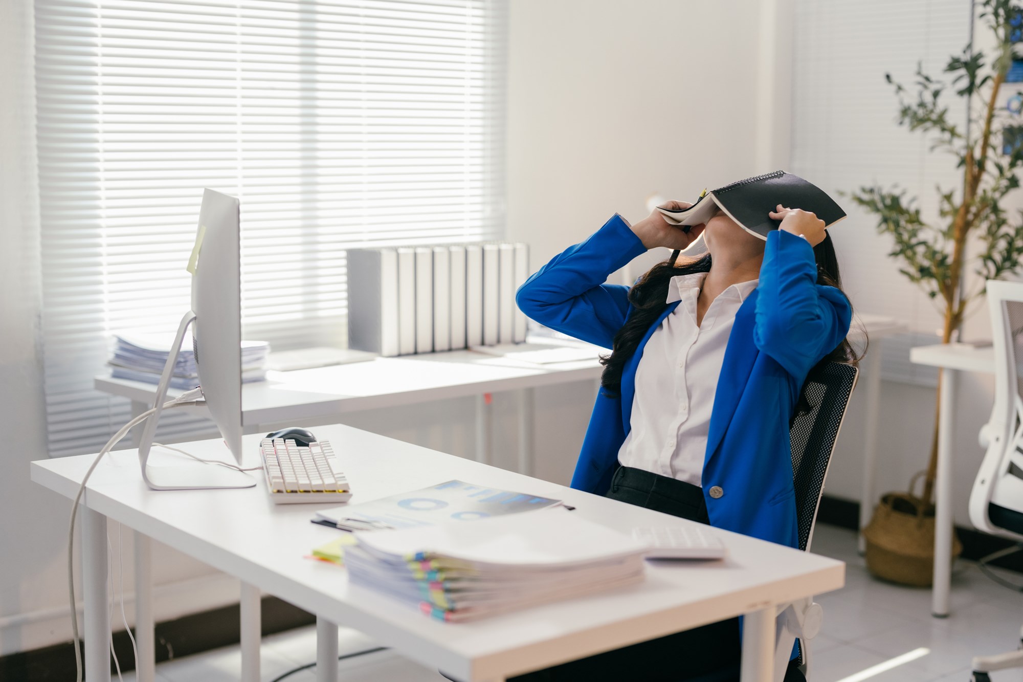 Stressed businesswoman is covering her face with a notebook while sitting at her desk in a modern office, overwhelmed by work and seeking a moment of peace