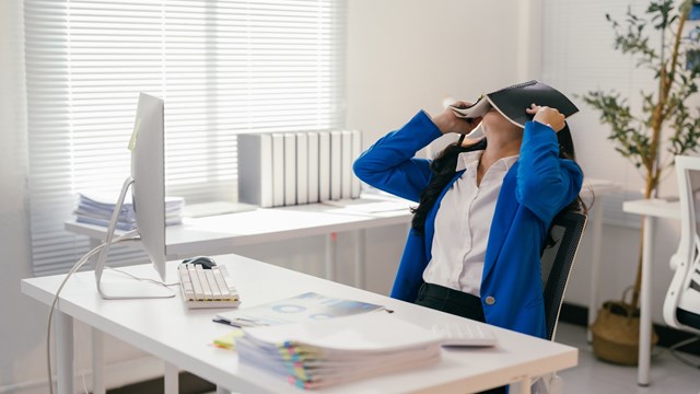 Stressed businesswoman is covering her face with a notebook while sitting at her desk in a modern office, overwhelmed by work and seeking a moment of peace