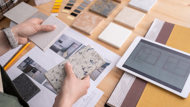 Hands of young female designer holding two samples of marble tile over wooden table with digital tablet, photos of home interior etc
