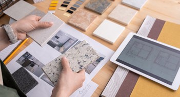 Common Area Design Hands of young female designer holding two samples of marble tile over wooden table with digital tablet, photos of home interior etc