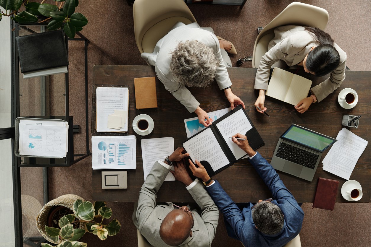 Group of middle aged multiethnic business professionals collaborating around table, reviewing documents and using laptop, top view showing teamwork and corporate meeting environment